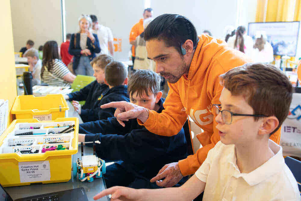 A teacher guiding students in a classroom, working with robotics kits and computers to learn coding and engineering.