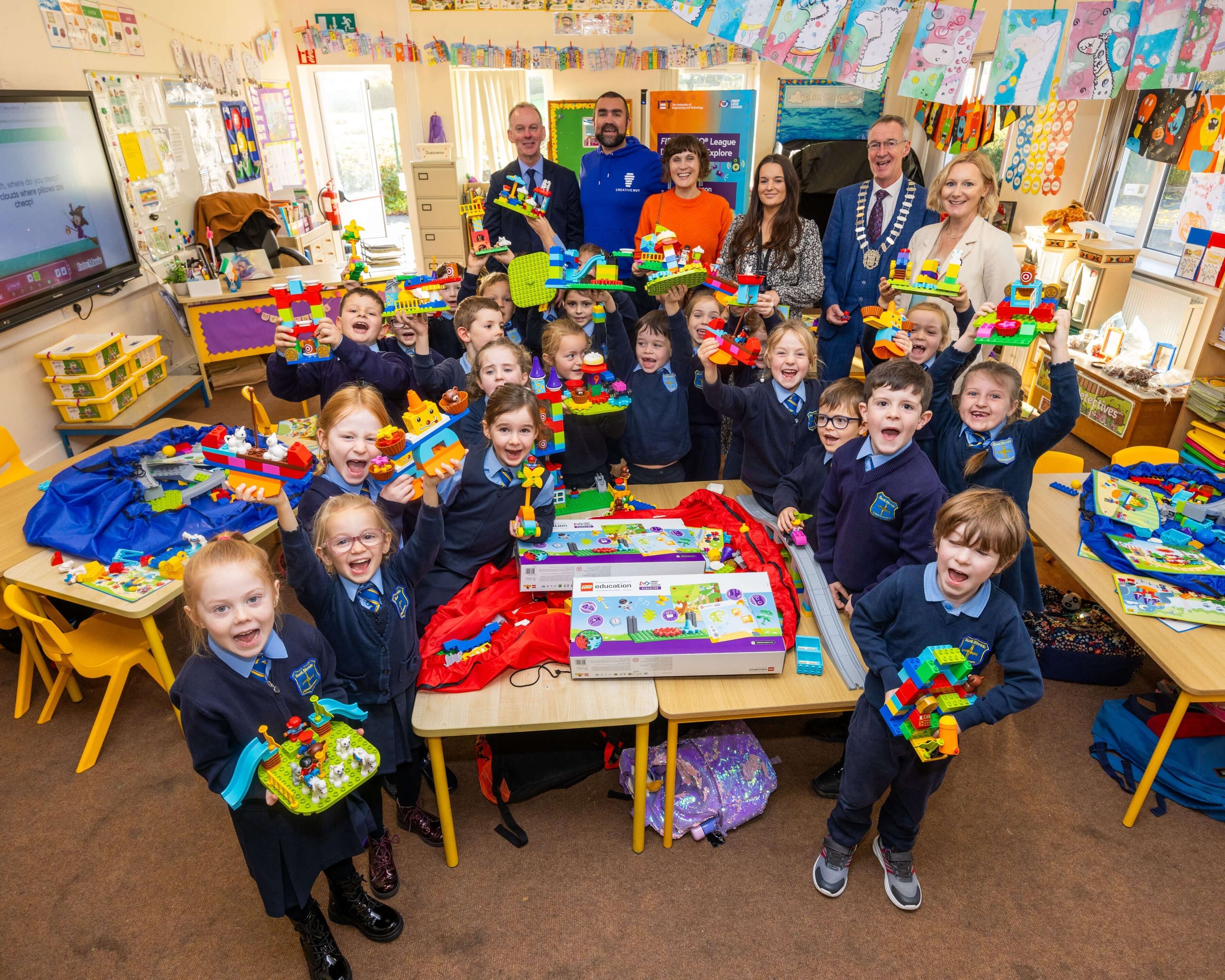 Students in a classroom proudly displaying their colorful building projects with teachers and guests.