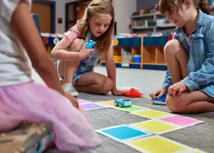 Three children engaged in a fun coding activity with toy cars on colorful markers in a classroom setting.