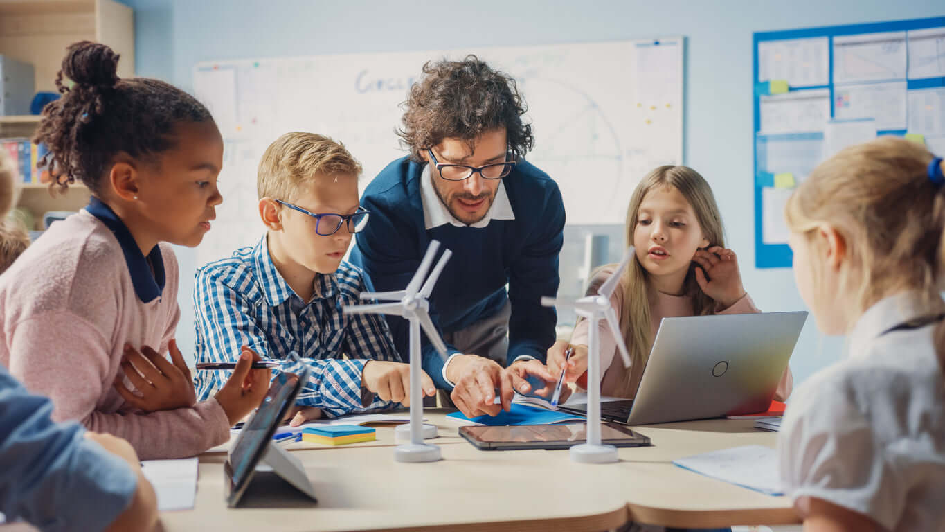 Teacher guides students in a hands-on science project with wind turbines and laptops in a classroom setting.