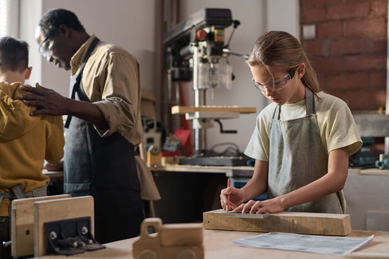 A young girl in safety goggles works on a woodworking project at a workshop with an instructor nearby.