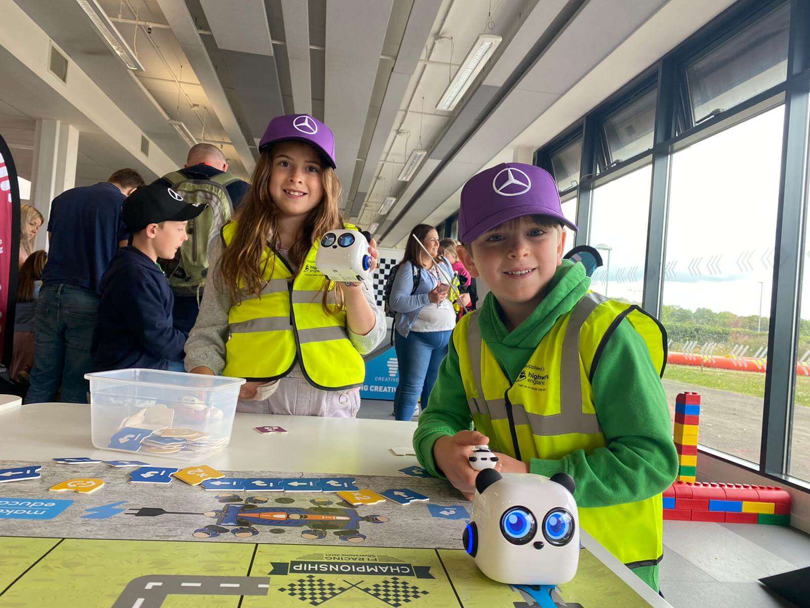 Two children in safety vests and caps play with robotic toys at a STEM event, engaged in hands-on learning.