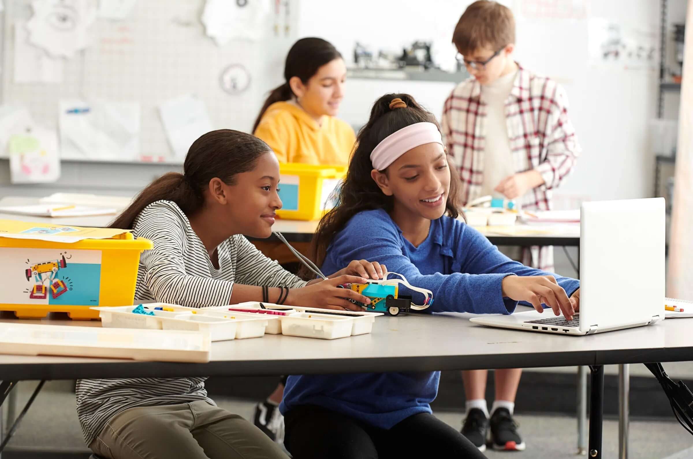 Two girls collaborating on a robotics project at a table, using a laptop and colorful building kits in a classroom setting.