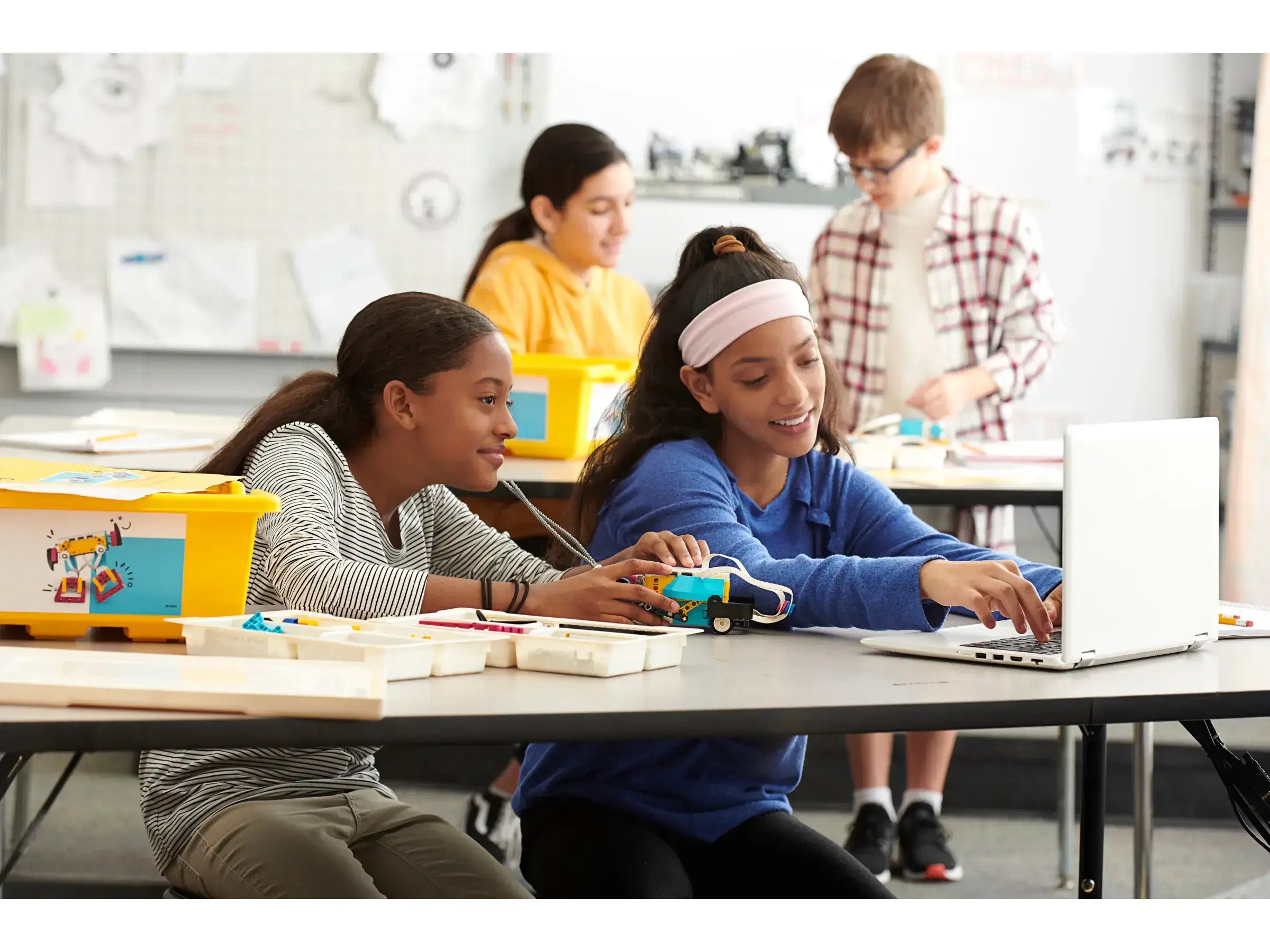 Two girls collaborate on a robotics project at a classroom table while a boy works on a laptop in the background.