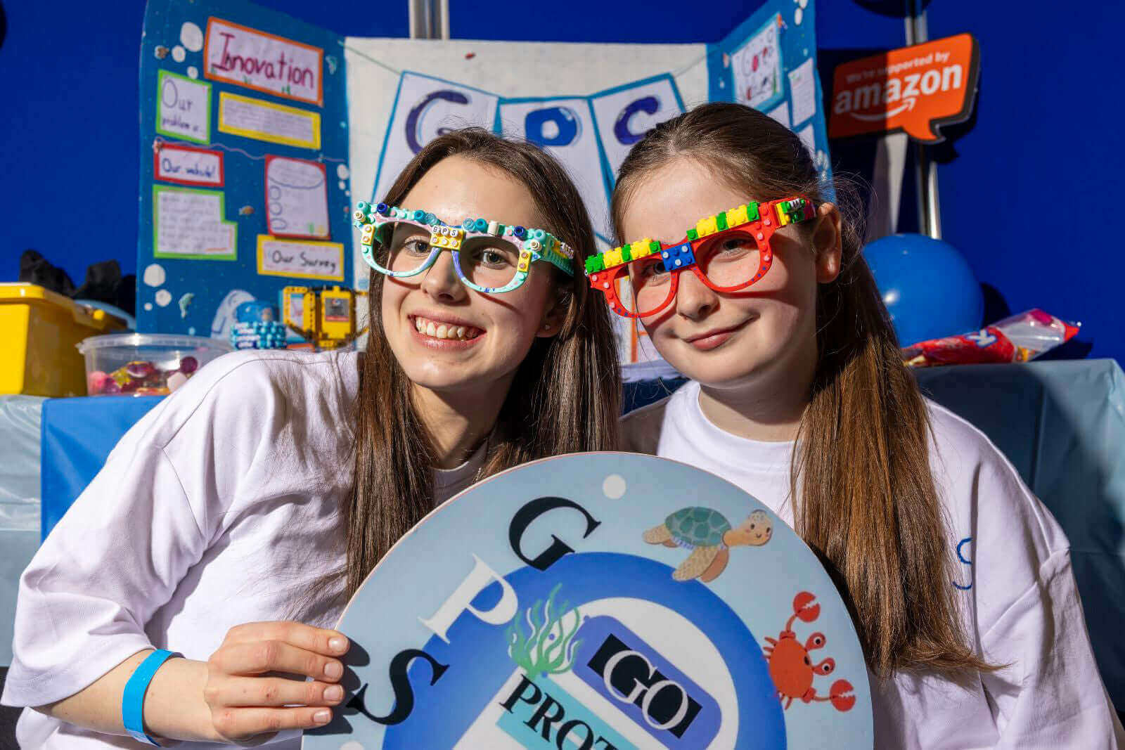 Two young girls wearing colorful glasses smile while holding a sign at a science fair booth focused on innovation.