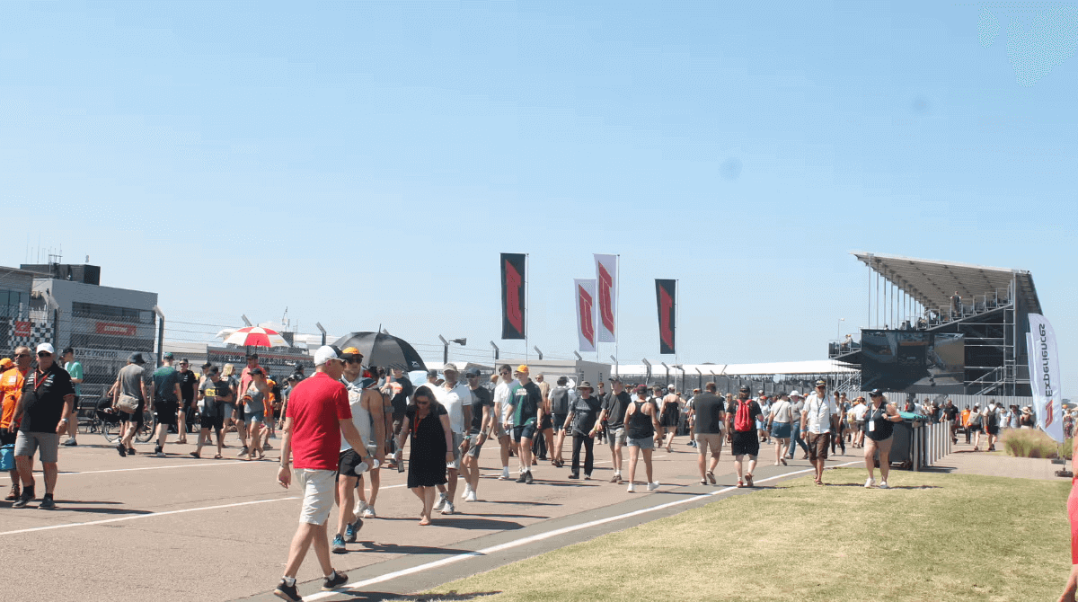 Crowd walking at an outdoor event with grandstands and flags in the background under clear blue skies.