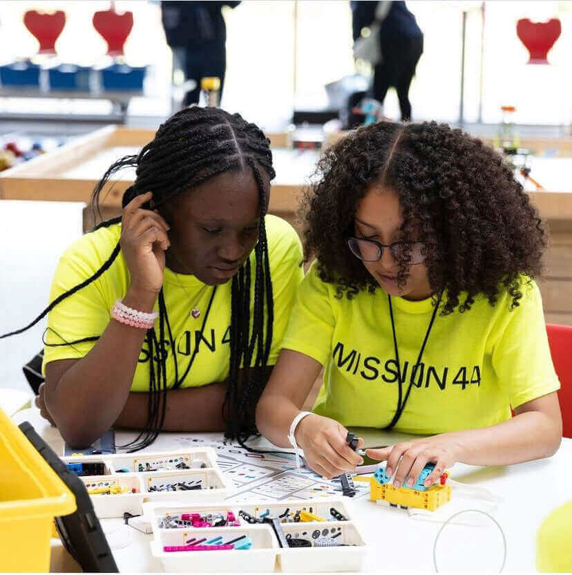 Two girls in bright yellow shirts engaged in a creative STEM project, working with colorful tools and materials.