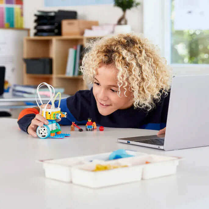 Young child exploring robotics with a colorful toy robot and laptop on a bright classroom table.