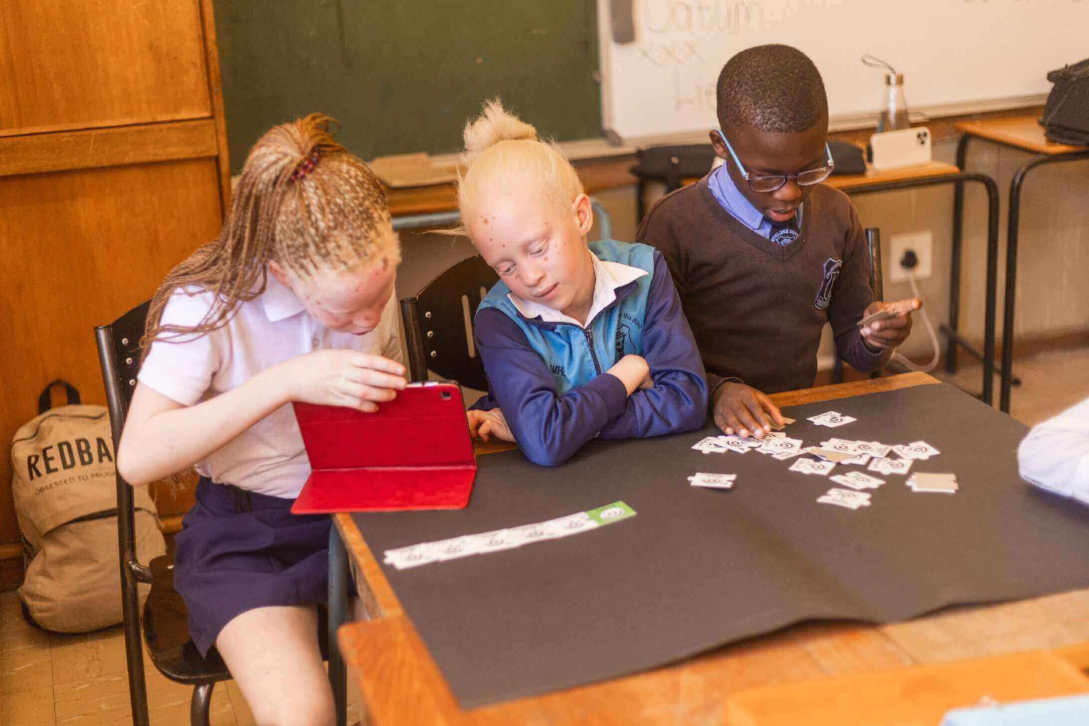 Children engaged in a creative activity in a classroom, using a tablet and playing with cards on a table.