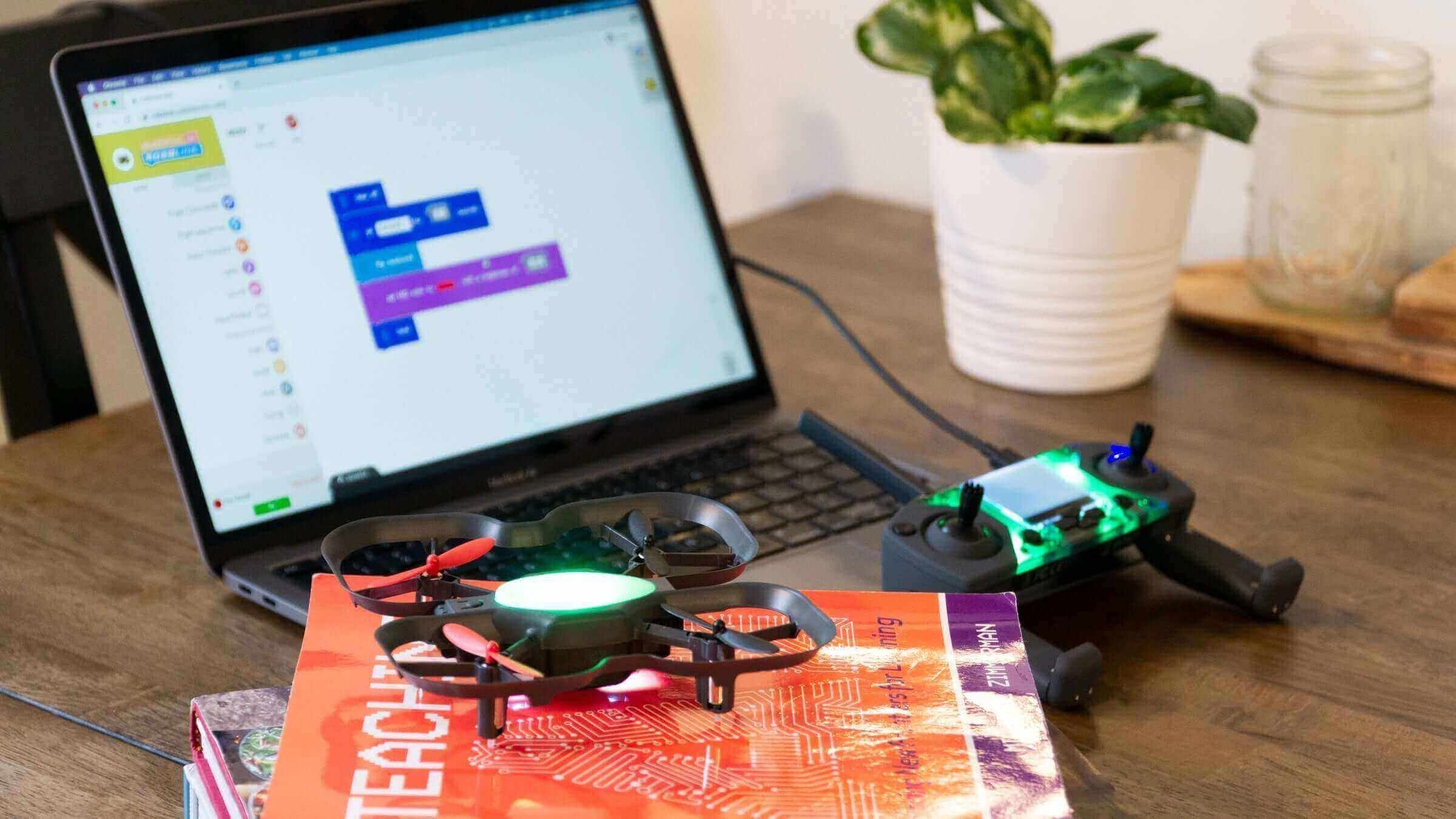 A laptop displaying coding software next to a drone and textbooks on a wooden table.
