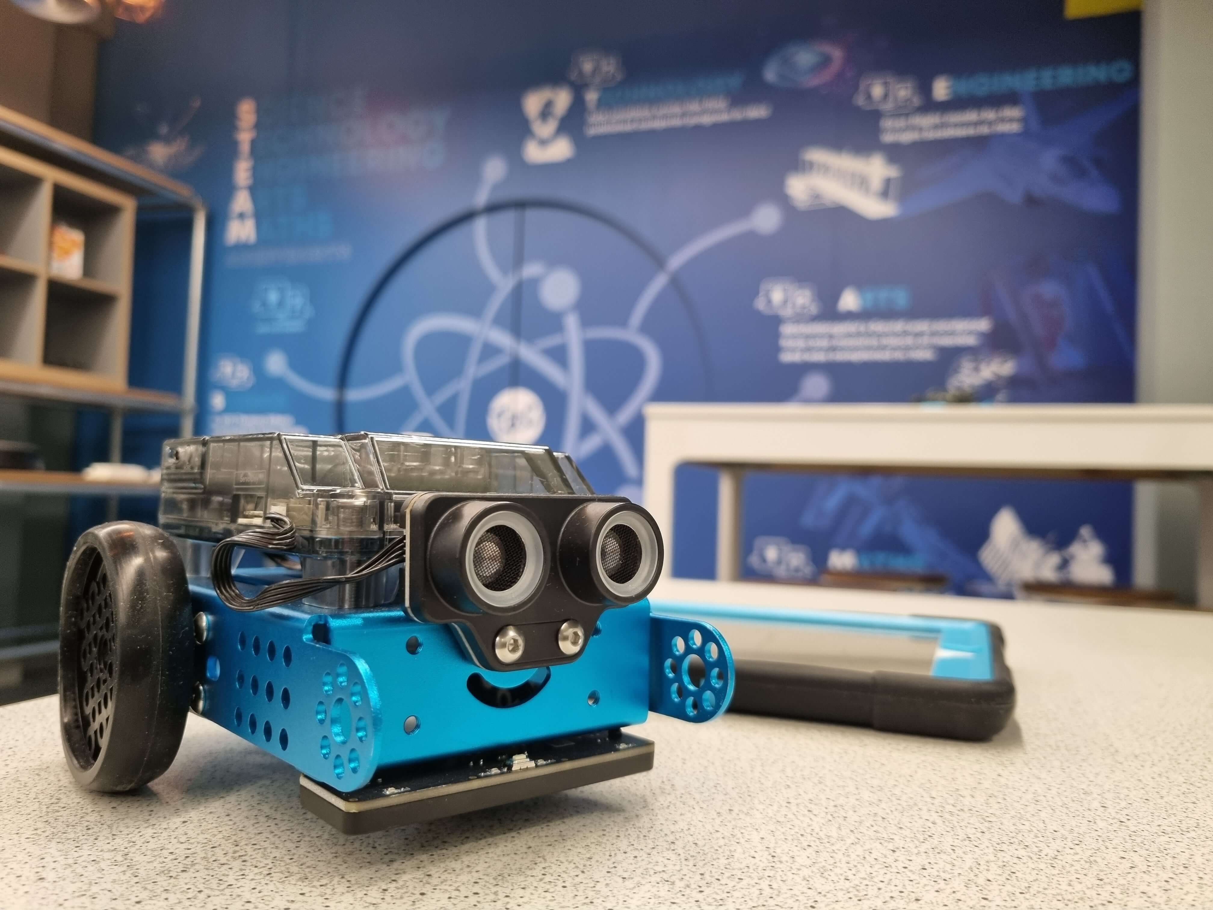A blue and black robotic toy car with a smiling face, placed on a table with educational posters in the background.