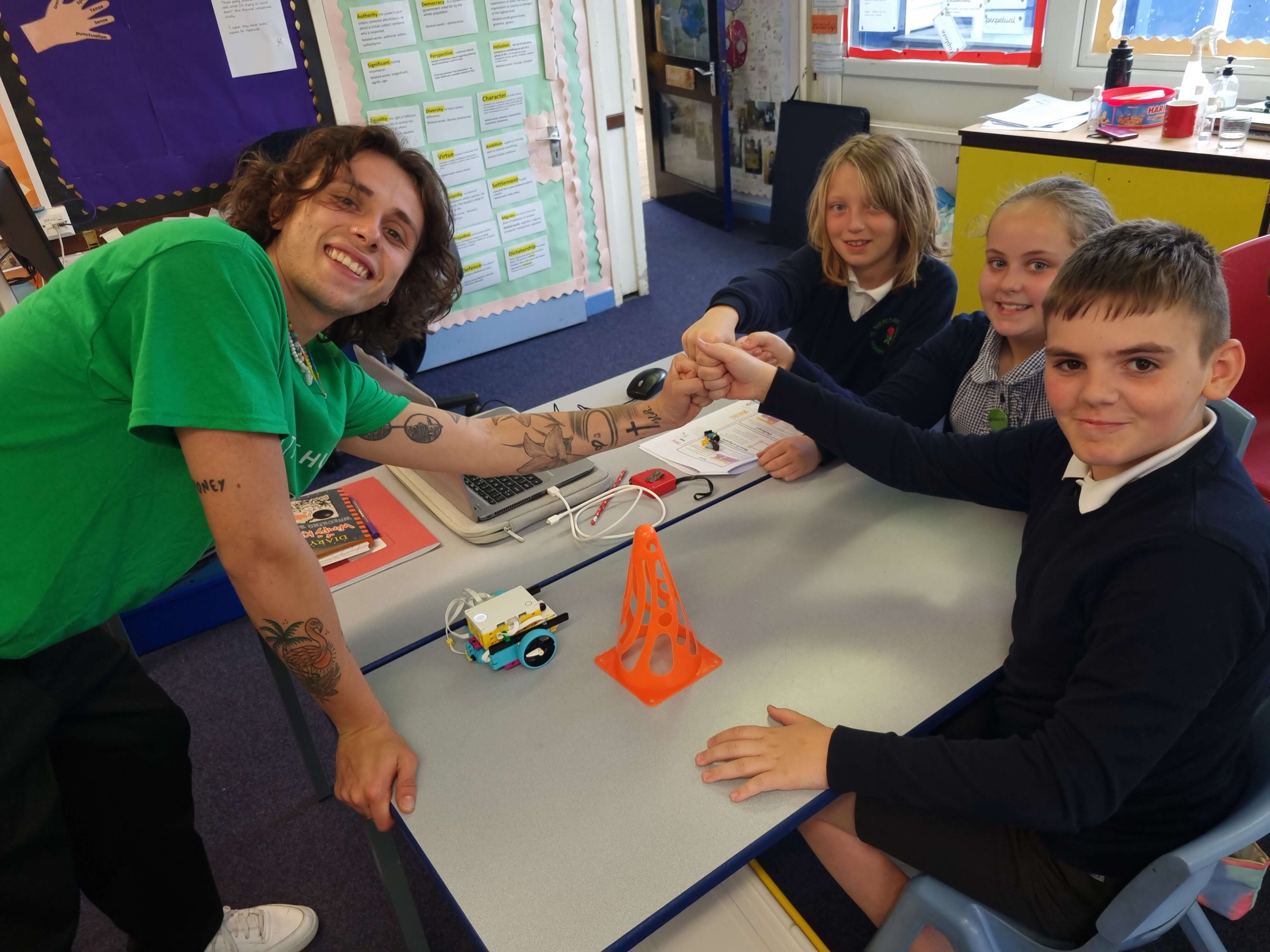 A teacher smiles with students, showcasing a robot on a table amid classroom learning activities.