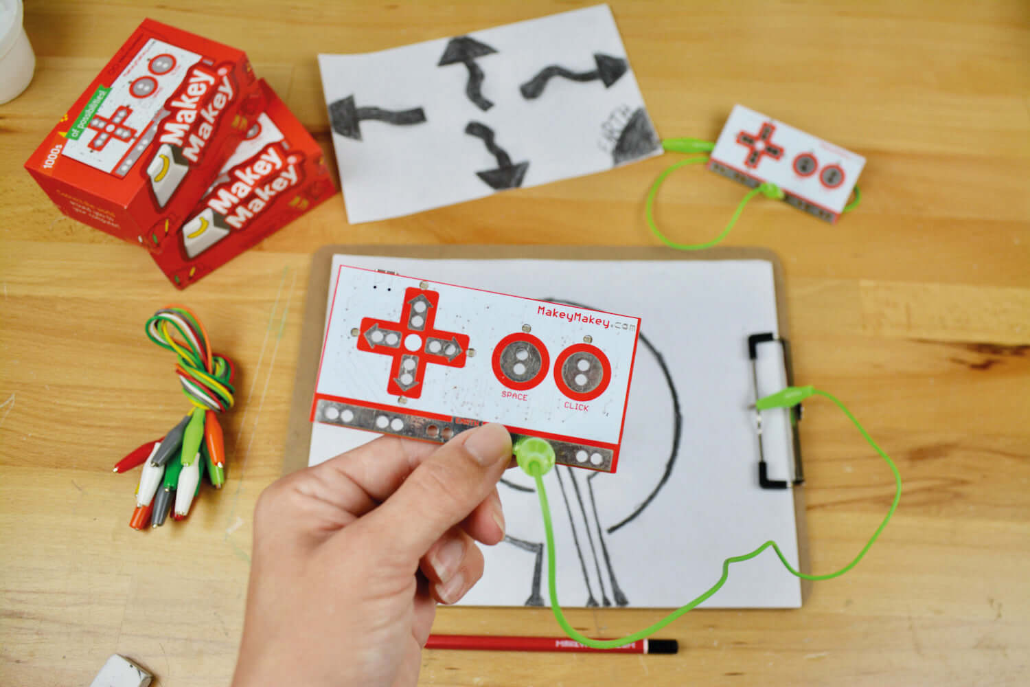 A person holding a Makey Makey circuit board with wires, alongside colorful markers and drawings on a wooden table.