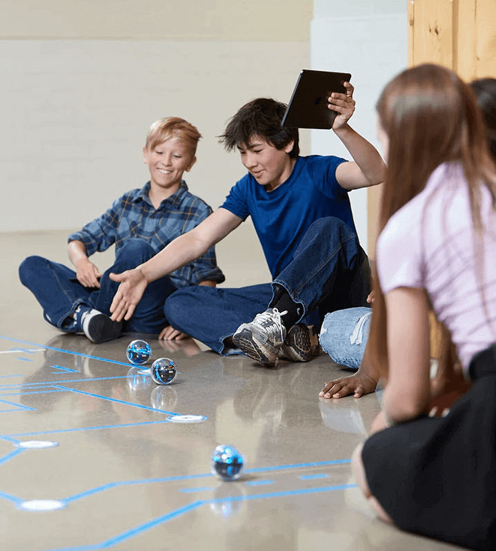 Children using a tablet to control robotic balls on the floor during a fun coding activity.