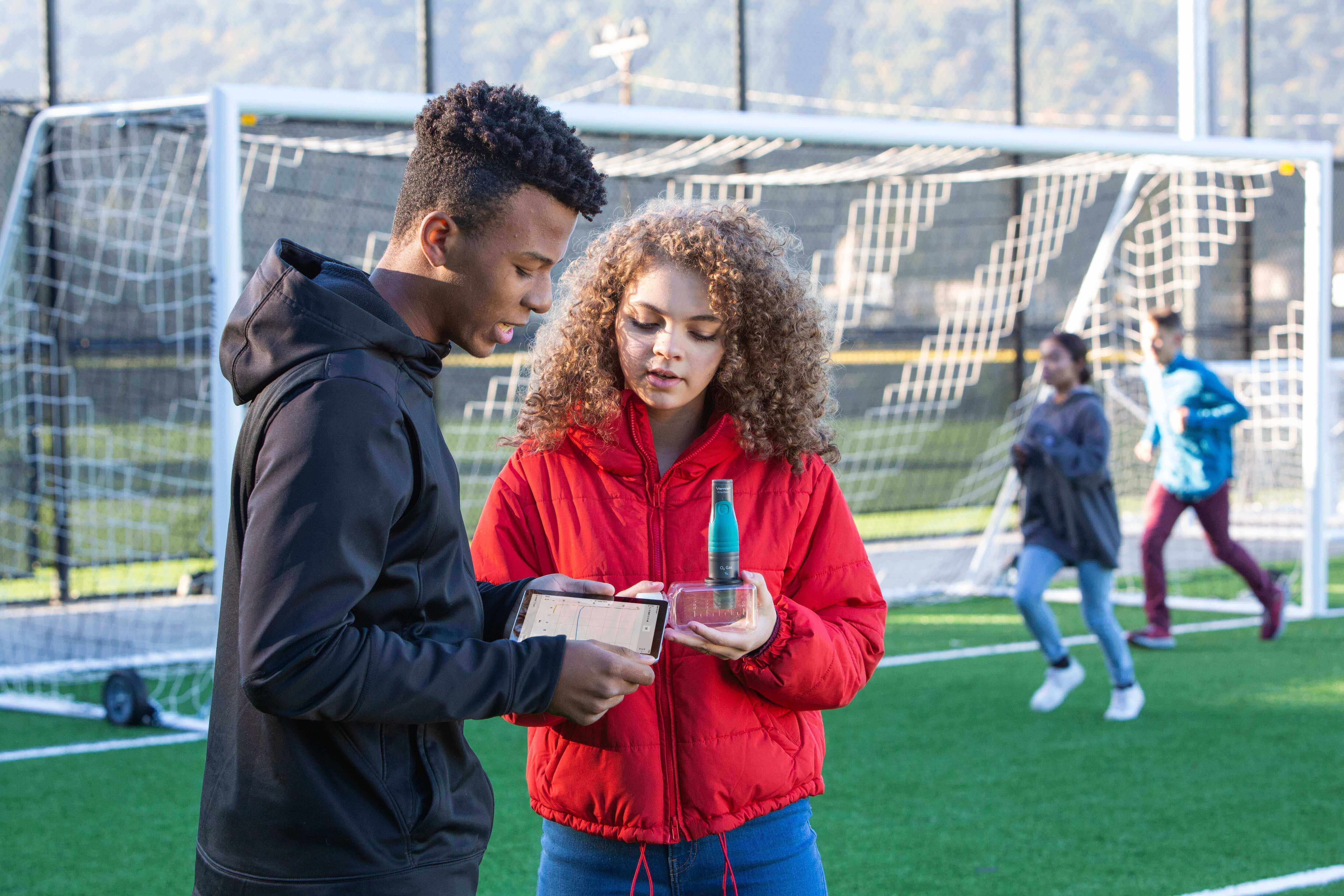 Two teenagers discussing a project on a soccer field, with a goal in the background and other kids playing in the distance.