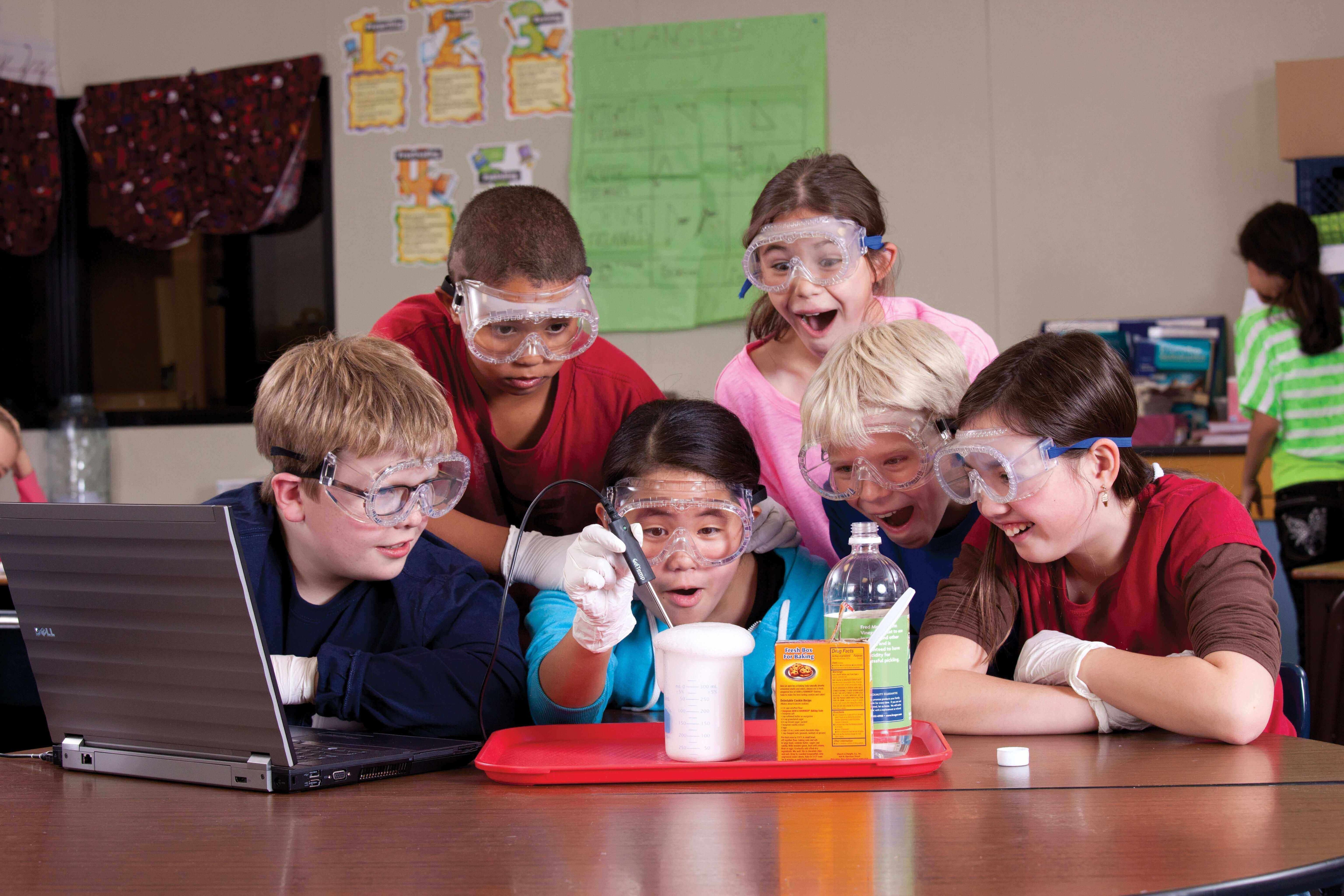 Excited children wearing goggles and gloves conducting a science experiment in a classroom, gathered around a laptop.