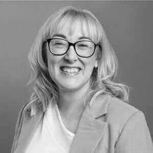 Smiling woman with blonde hair and glasses wearing a light blazer, set against a neutral background.
