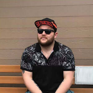Man in sunglasses and a baseball cap sitting on a bench, wearing a patterned shirt.