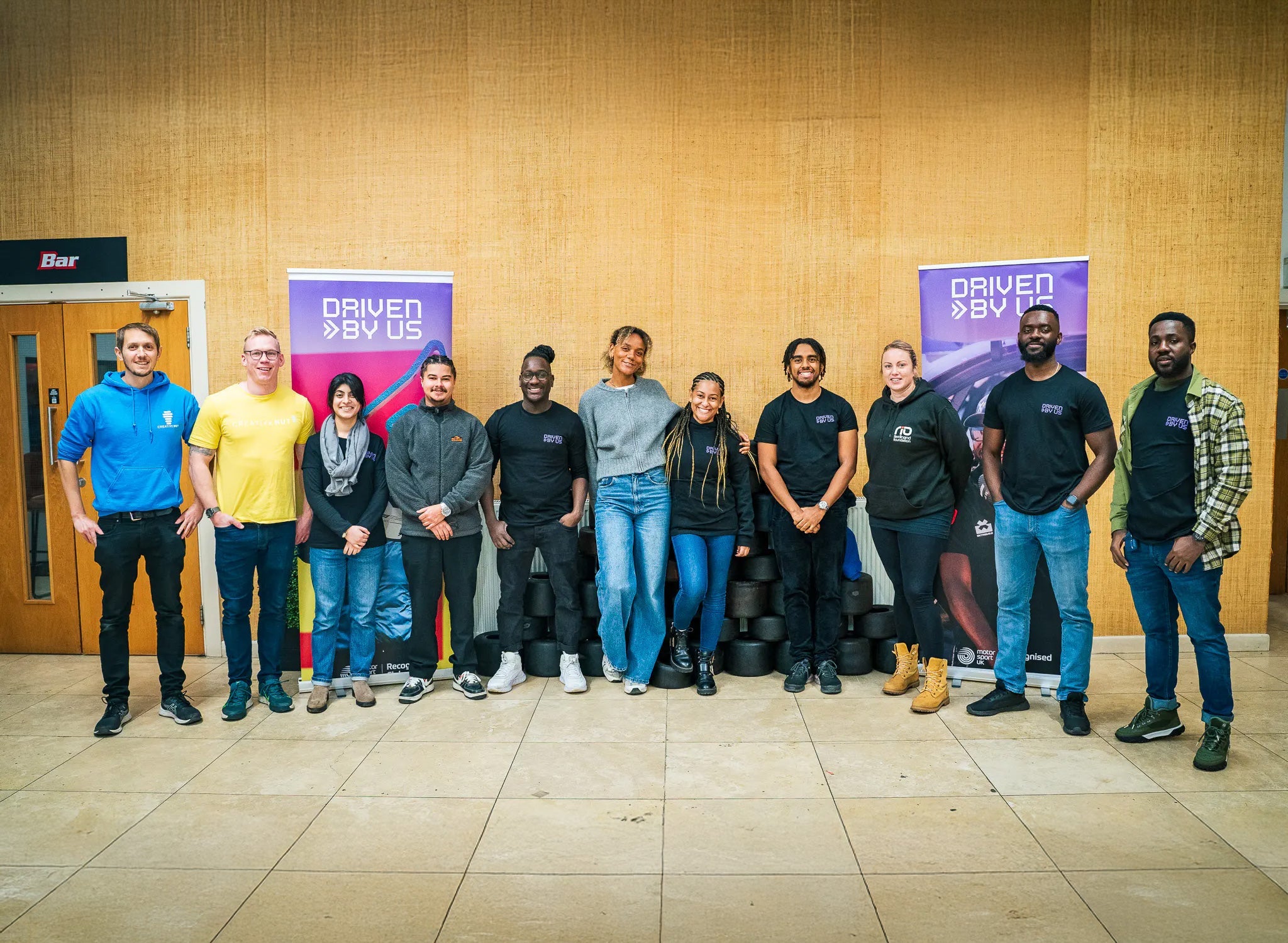Group photo of diverse individuals at Driven By Us event, standing in front of promotional banners and tires.