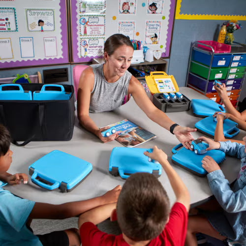 Teacher engaging with children at a classroom table, distributing blue educational kits while fostering collaboration and learning.