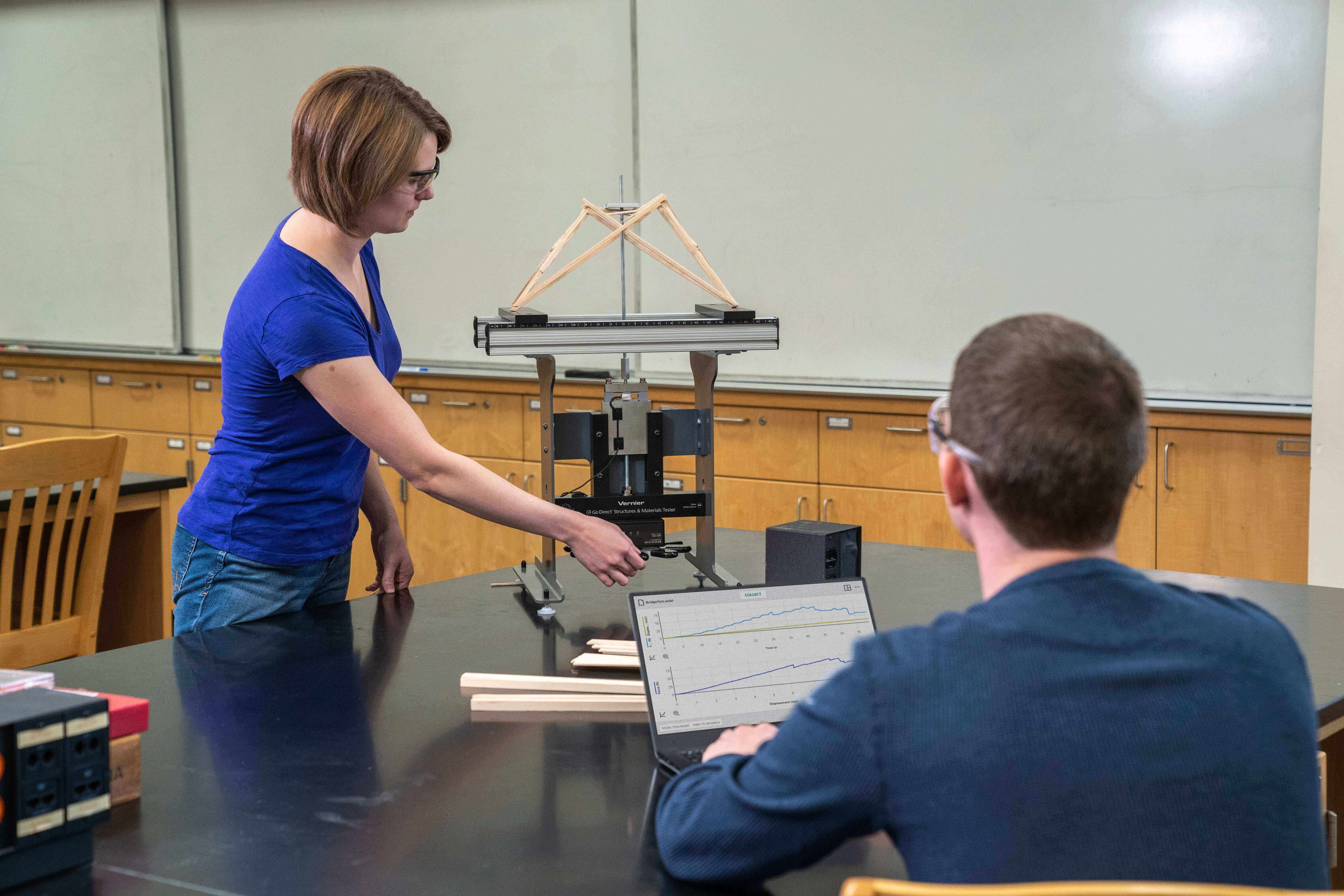 Student demonstrating a physics experiment using a testing machine and laptop in a classroom lab.