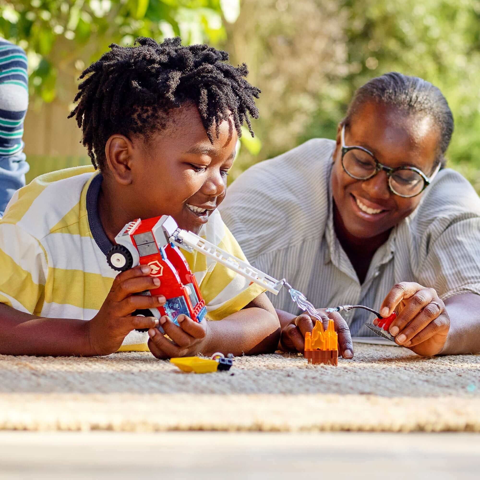 Child playing with LEGO® City Fire Ladder Truck, enjoying quality time with a family member in a sunny, cheerful environment.
