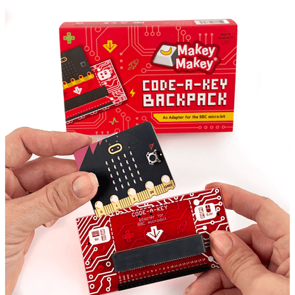 Hands holding Makey Makey Code-a-Key Backpack adapter for BBC micro:bit with packaging in the background.