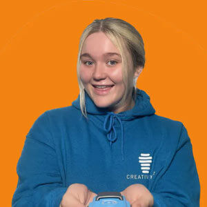 Smiling young woman in a blue hoodie holding a device, against an orange background, promoting creativity.