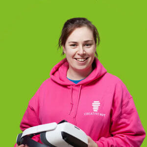 Smiling woman in a pink hoodie holding a VR headset against a bright green background.